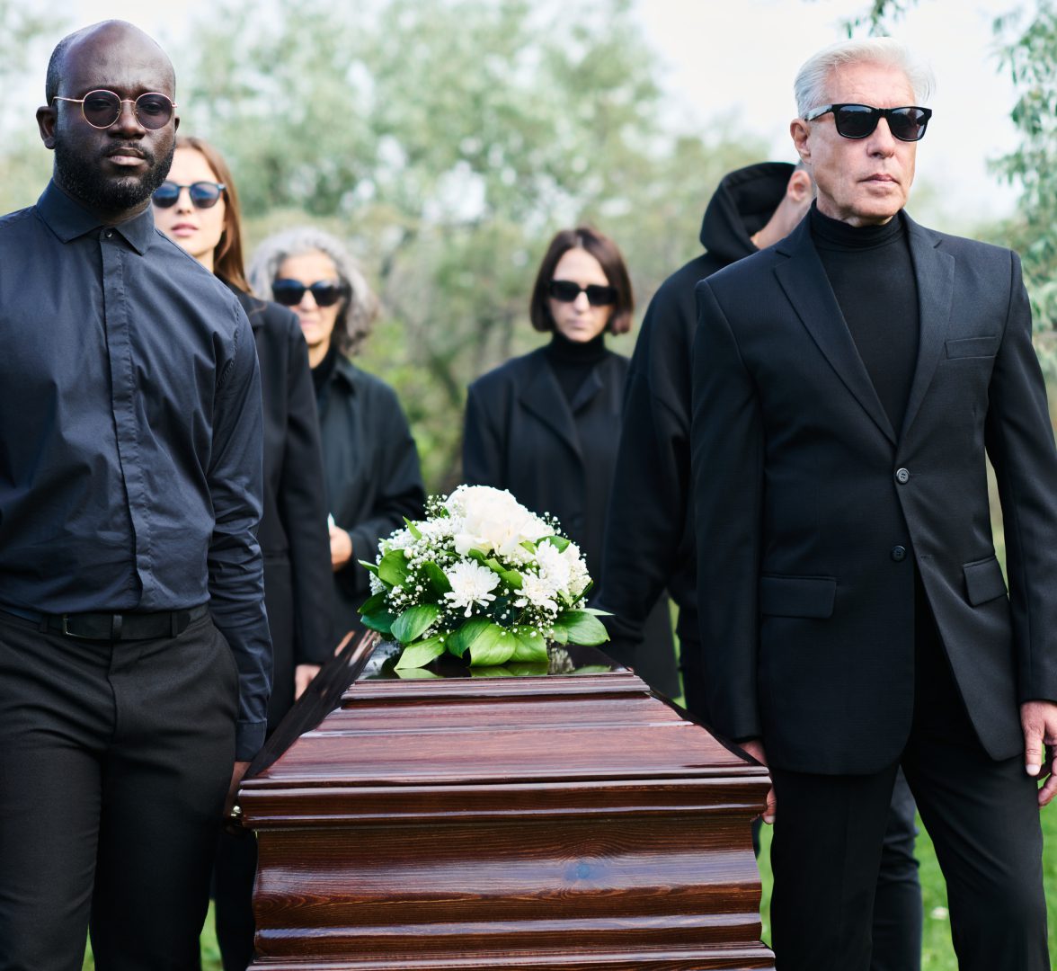 People dressed in black suits and sunglasses carry a wooden casket adorned with white flowers in an outdoor setting.
