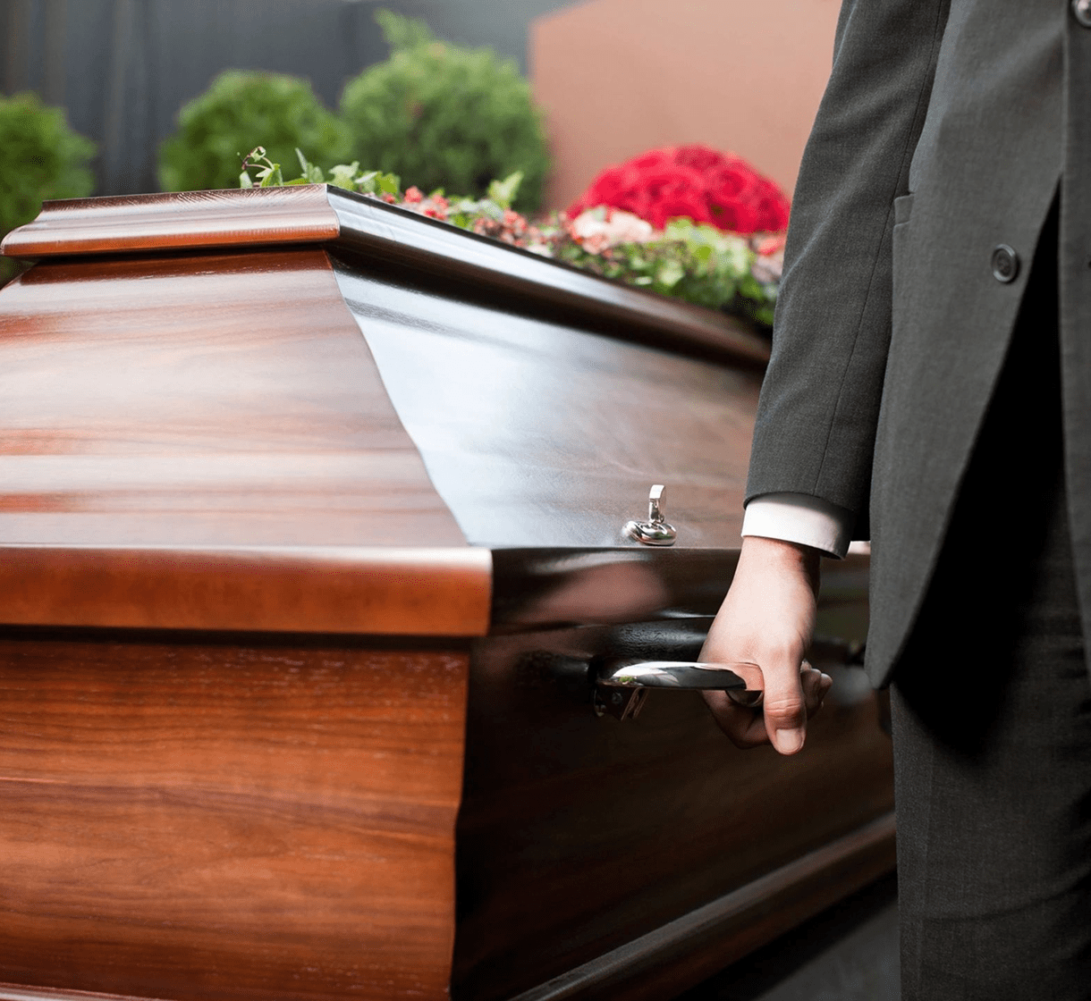 Person in a black suit holding the handle of a wooden casket adorned with flowers.