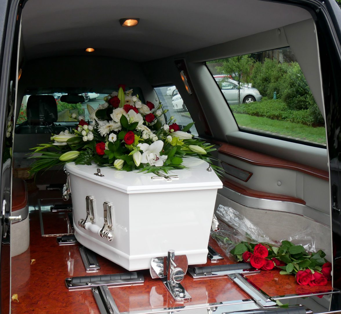 A white coffin adorned with red and white flowers is placed inside a hearse, with additional red roses nearby.