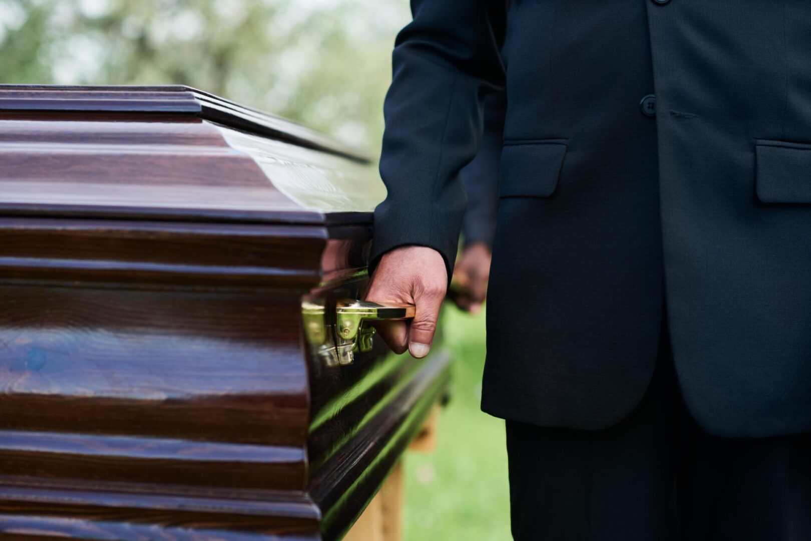 Person in black suit holding a handle of a wooden coffin outside.