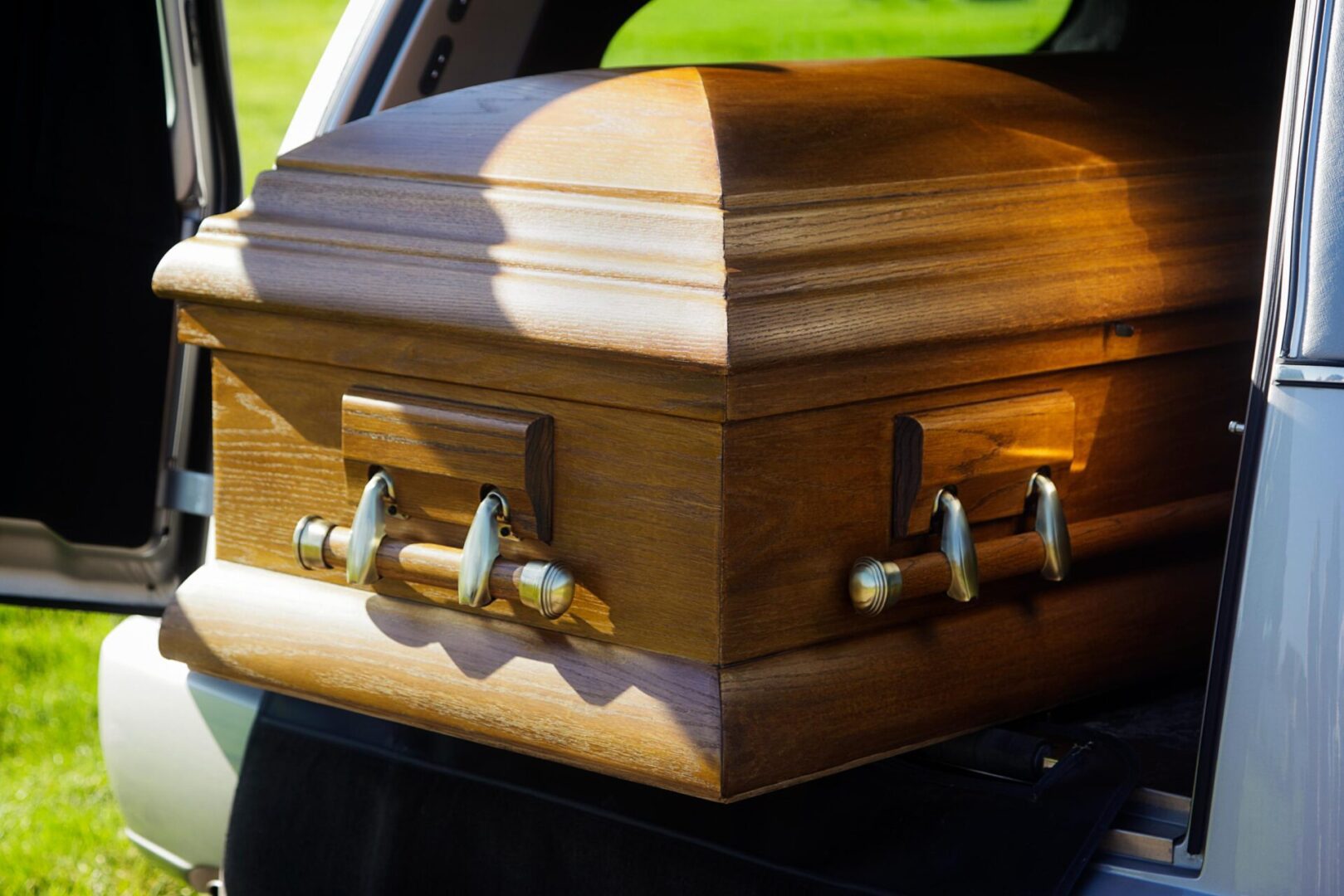 A wooden coffin partially extended from the back of a hearse in daylight.