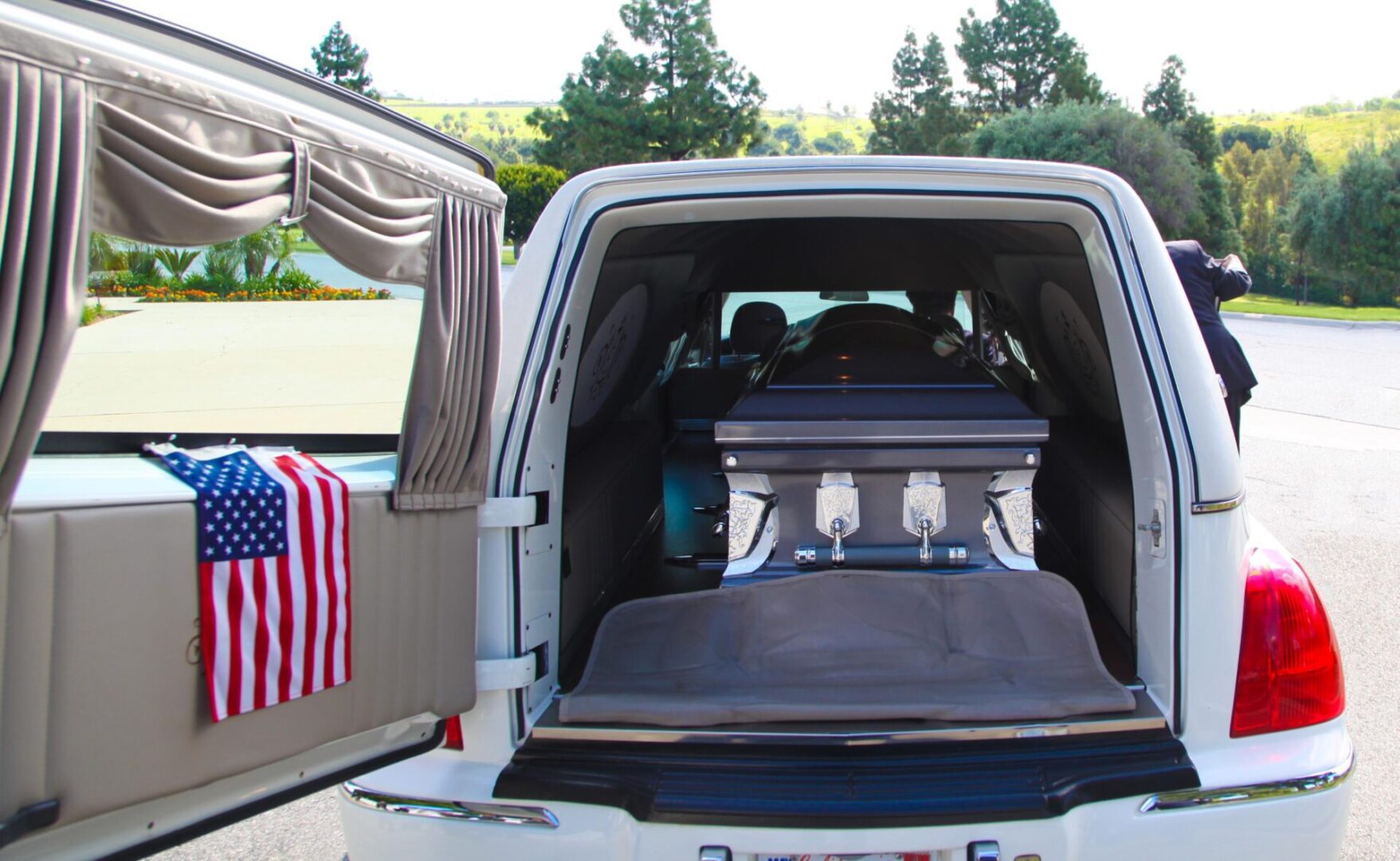 A hearse with an open rear door reveals a casket inside. An American flag is displayed on the left door. Trees and a road are visible in the background.