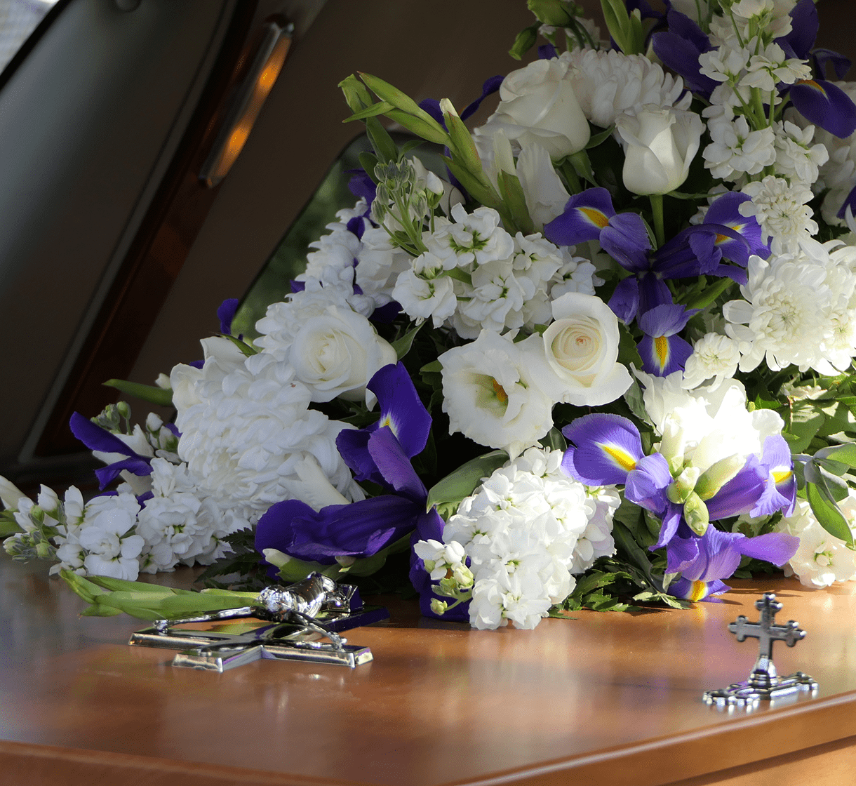 A bouquet of white roses and purple irises rests on a polished wooden casket, with two metal crosses placed beside it.