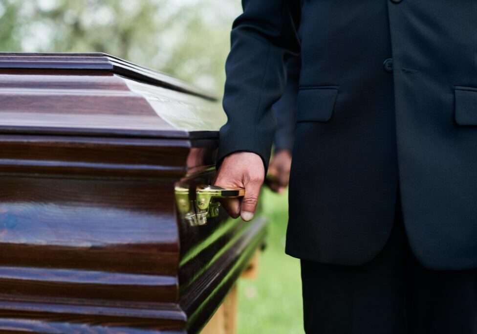 Person in black suit holding a handle of a wooden coffin outside.