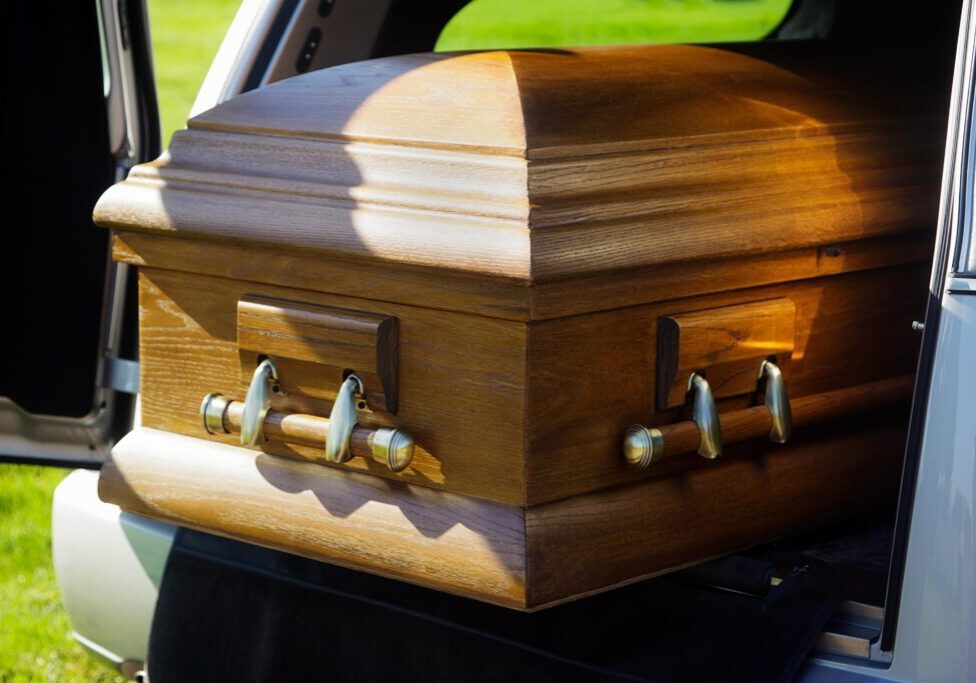 A wooden coffin partially extended from the back of a hearse in daylight.