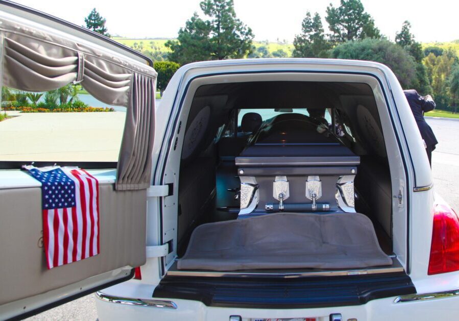 A hearse with an open rear door reveals a casket inside. An American flag is displayed on the left door. Trees and a road are visible in the background.